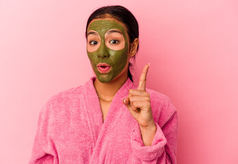 Young Venezuelan woman wearing a bathrobe and facial mask isolated on pink background having an idea, inspiration concept.