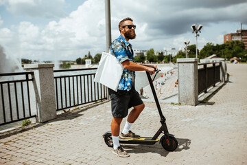 Young hipster man driving an electric scooter on a bright sunny day holding white textile eco bag...