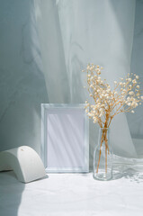 Empty picture frame on gray. Interior mock up design concept with shadows and sunlight. White blank wooden frame mockup, gypsophila flowers in glass vase, geometric shape on the table.