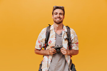 Smiling tourist with ginger beard in short sleeve summer shirt and plaid t-shirt holding camera and smiling on orange backdrop.. - obrazy, fototapety, plakaty