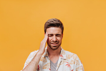 Trendy man with stylish beard in white summer printed shirt and grey t-shirt with headache posing with closed eyes on orange backdrop..
