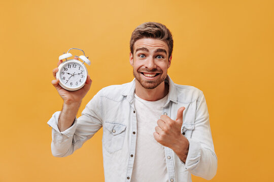 Smiling blue eyed stylish man with dinger beard in white t-shirt and denim cool jacket looking into camera and posing with big alarm clock..