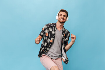 Fashionable cool man with beard in grey t-shirt, pineapple and palm print shirt and shorts smiling, looking into camera and posing with headphones..