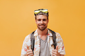 Joyful bearded tourist with green swimming glasses in white printed outfit looking into camera on orange isolated background..