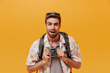 Bearded man with blue eyes and stylish hairstyle in white printed outfit and backpack posing with camera on orange background..