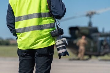 A military photographer, a reporter, in a signal yellow vest, ready to work in front of a military helicopter. Close-up. The background is blurred.