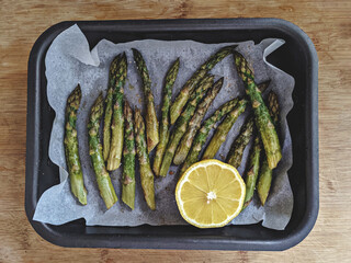 Baked asparagus in baking dish cooking in oven and half of lemon on wooden surface table, healthy...