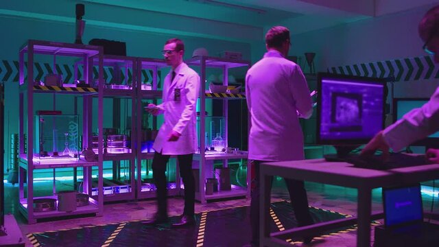 Three professional male scientists in white coats working inside modern futuristic laboratory . Man looking at computer screen  doing experiment . Physicians walking inside room . Scientific equipment