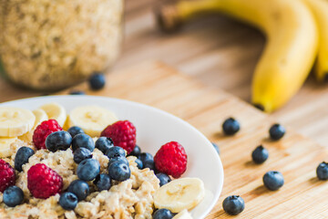 Porridge with fresh raspberries, blueberries and banana slices on a white plate. Nutritious and delicious breakfast. - obrazy, fototapety, plakaty