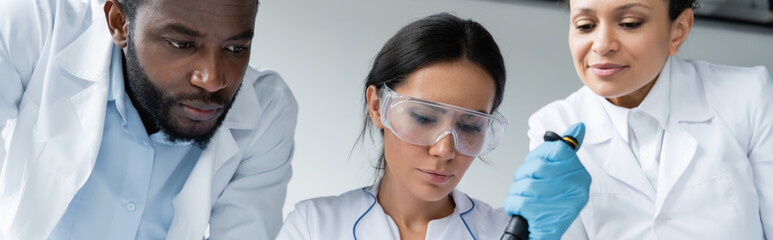 Scientist in goggles holding pipette near interracial colleagues, banner