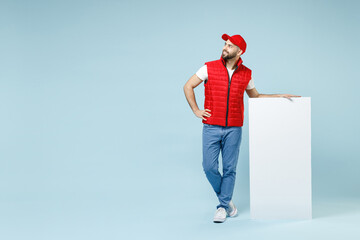 Full size body length delivery guy employee man in red cap white T-shirt vest uniform work as courier hold big blank poster promo area isolated on pastel blue color background studio. Service concept.