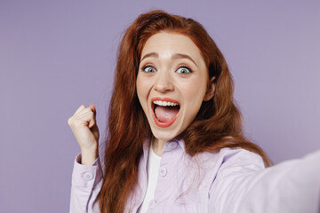 Close up excited young redhead curly woman 20s wears white T-shirt violet jacket doing selfie shot on mobile phone clench fists say yes isolated on pastel purple color wall background studio portrait