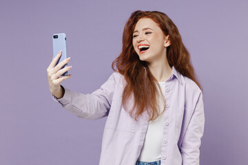 Smiling overjoyed fun young redhead curly green-eyed woman 20s wears white T-shirt violet jacket doing selfie shot on mobile cell phone isolated on pastel purple color wall background studio portrait