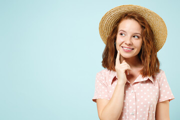 Smiling happy lost in thought young redhead woman puts finger on cheek wears casual pink dress straw hat look aside isolated on pastel blue color background studio portrait. People emotions concept