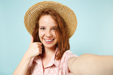 Smiling happy young redhead woman 20s doing selfie shot on mobile phone showing thumb up gesture wear casual pink dress straw hat look camera isolated on pastel blue color background studio portrait