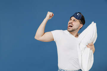 Naklejka premium Young stressed threatening annoyed sad tired man in pajama jam sleep mask rest relax at home hold pillow scream look aside clench fist isolated on dark blue background Bad mood night bedtime concept
