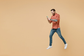 Fototapeta na wymiar Full size body length satisfied smiling fun tatooed young brunet man 20s short haircut wears apricot shirt jump hold in hand use mobile cell phone isolated on pastel orange background studio portrait