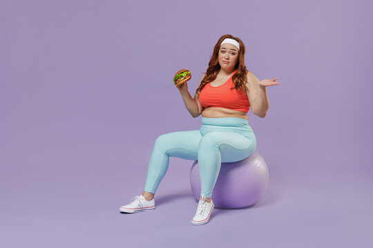 Full length addicted young chubby overweight plus size big fat woman in top warm up train sit on fit ball biting fast food burger spread hand isolated on purple background gym Workout sport concept
