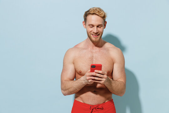 Handsome young sexy blond smiling fun man 20s in red shorts swimsuit relax near hotel pool hold using mobile cell phone isolated on pastel blue background. Summer vacation sea rest sun tan concept