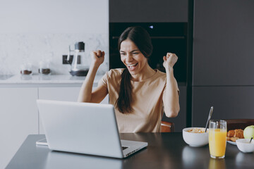 Young excited fun housewife woman 20s in beige shirt eat breakfast using laptop pc computer look for recipe do winner gesture cook food in light kitchen at home alone. Healthy diet lifestyle concept.