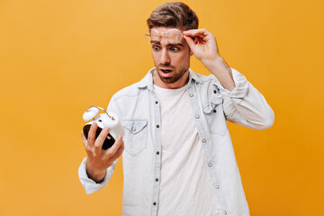 Surprised man with brown hair in clear round eyeglasses, denim shirt and white t-shirt looking at alarm clock on isolated backdrop..