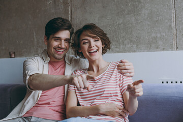 Young surprised fun couple two friends woman man 20s in striped t-shirt casual clothes sitting on sofa hugs watch tv point index finger resting indoors at home flat together People lifestyle concept.