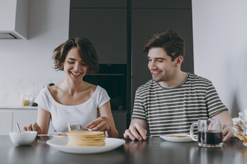 Young smiling fun happy couple two woman man in casual t-shirt clothes sit by table eat pancakes with maple syrup in morning cook food in light kitchen at home together Healthy diet lifestyle concept.