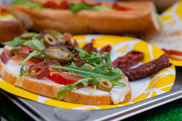 Sandwich with arugula, ham, cheese, jalapeno, sundried tomato on plate on counter at summer local food market - close up. Gastronomy, street food, mediterranean and Italian dish concept