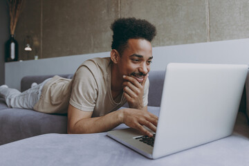 Young smiling fun freelancer african american man 20s wear beige t-shirt sweatpants lay down on grey sofa indoors apartment use laptop pc computer chatting resting on quarantine weekends stay home - obrazy, fototapety, plakaty