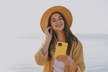 Young satisfied woman in straw hat shirt summer casual clothes earphones use mobile cell phone listen music outdoors at sunrise sun dawn over sea background People vacation lifestyle journey concept
