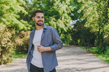 Young satisfied smiling happy man in blue casual shirt t-shirt walking look aside rest relax in spring green city park go down alley sunshine lawn outdoors on nature Urban lifestyle leisure concept.