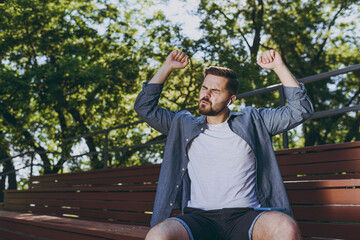 Bottom view happy young man in blue shirt shorts earphones sit on bench listen to music dance sing...