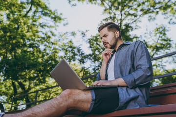 Bottom side view programmer young man in blue shirt shorts earphones sit on bench work online laptop pc computer wave hand rest relax in spring green city park outdoor on nature Urban leisure concept.