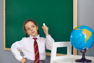 Thoughtful little girl with chalk standing in front of blackboard at classroom. Child having...