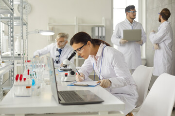 Obraz premium Young woman lab worker working with microscope making notes on clipboard. Scientist team group working at modern biochemistry medical laboratory. Scientific research on drugs and vaccine development