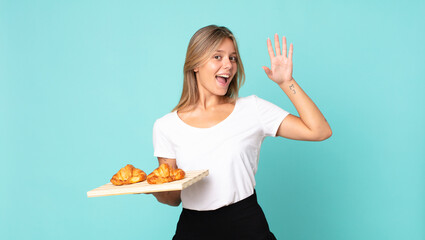 young blonde woman smiling happily, waving hand, welcoming and greeting you and holding a croissant tray
