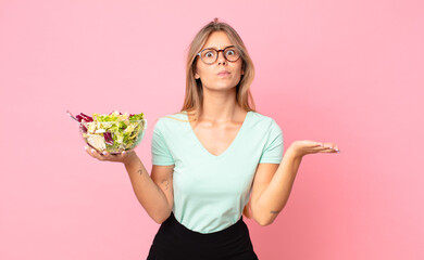 young blonde woman feeling puzzled and confused and doubting and holding a salad - obrazy, fototapety, plakaty