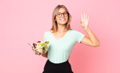 young blonde woman smiling happily, waving hand, welcoming and greeting you and holding a salad