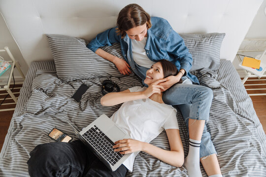 An Overhead View Of The Positive Lesbian Couple With Laptop Holding Hands And Looking To Each Other While Lying On The Bed