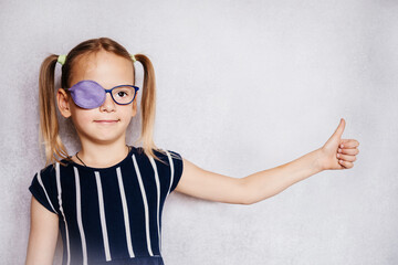Little girl wearing glasses and eye patch or occluder with her thumb up, amblyopia (lazy eye)...
