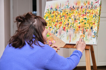 Young woman painting on canvas with oil paints at home. Artist drawing on easel sitting on the floor.