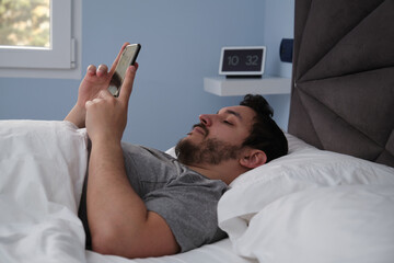 Young latin man with his smartphone in bed late in the morning. Digital clock at the background.