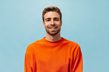 Positive young guy with red beard in bright orange sweatshirt looking into camera and smiling on...