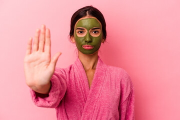 Young caucasian woman wearing a bathrobe and facial mask isolated on pink background standing with outstretched hand showing stop sign, preventing you.