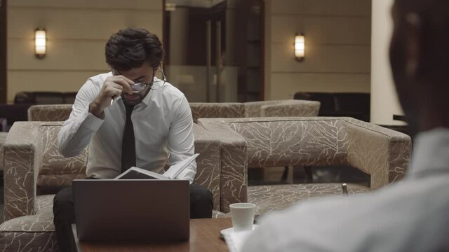 Over shoulder of unrecognizable contractor having private meeting with tired dark-haired entrepreneur sitting in armchair, putting down notebook, taking off glasses, talking