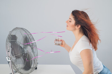 A red-haired Caucasian woman chills by the electric fan and drinks a cold drink. Climate control in...