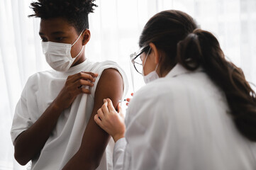 A female doctor or nurse wearing a mask and face shield is injecting the coronavirus 19 vaccine on the shoulder of an African-American man to immunize. Concept of preventing the spread of COVID-19.