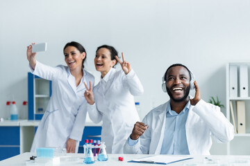 Cheerful african american scientist in headphones near colleagues taking selfie in lab