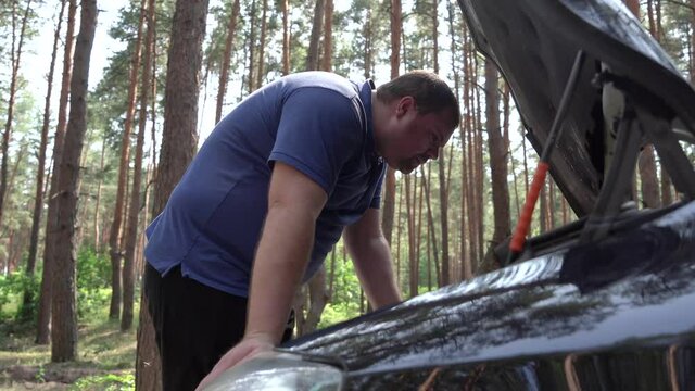 The car broke down in the woods and the man opens the hood to see what is wrong