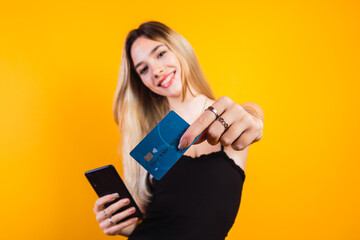 Portrait of a happy young blonde woman showing a plastic credit card while holding a cell phone isolated on yellow background.  Black friday.